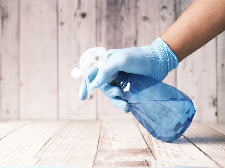 A gloved hand holding an unlabelled spray bottle over a pale wooden floor